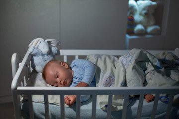 Adorable newborn baby boy, sleeping in crib at night. Little boy in white pajamas taking a nap in dark room. Bedroom interi?r with lamp and teddy bear . © Tomsickova