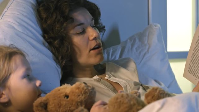 Medium shot of young woman reading aloud to cute little girl, who is hugging teddy bear and listening to bedtime story before going to sleep