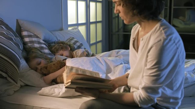 Medium shot of young mother reading bedtime story to two little kids of elementary school age, then turning off light and going away