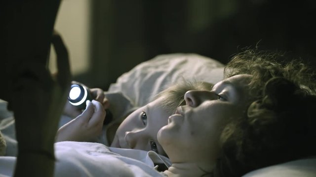 Tracking shot of caring young mother reading fairy tales to little girl, who is holding flashlight and lying on pillow next to her