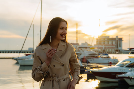 beautiful long-haired smiling girl in trendy beige coat posing in seaport at sunset