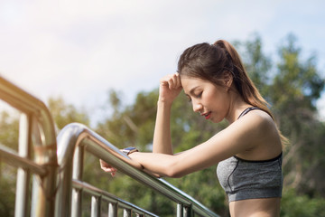 Young woman relaxing after jogging exercise on fence at park to freshen her body and enjoy warm light in morning. Young asian woman take a break from running execise. Outdoor exercise activity.