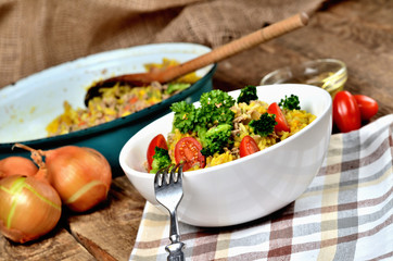 Fork and tuna risotto with vegetables, tomatoes, broccoli and parsley in the bowl, onions, oil and pan in the background