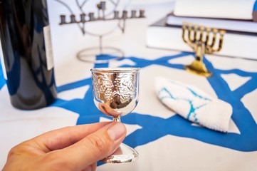 Hand holding a "kiddush" wine blessing cup, a traditional Jewish religious symbol. With more Jewish symbols on the background: prayer books, "menorah" lampstand on a flag of Israel. 