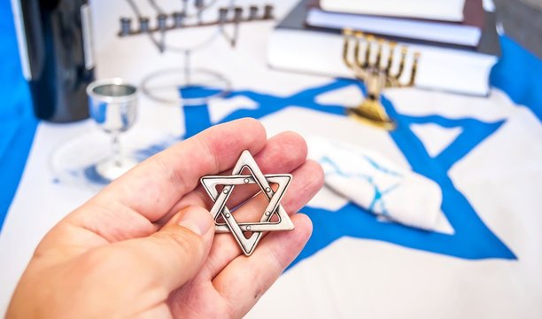Hand holding a David Star ("Magen David" in Hebrew), a traditional Jewish religious symbol. With more Jewish symbols on the background: prayer books, menorah lampstand, kiddush wine blessing glass 