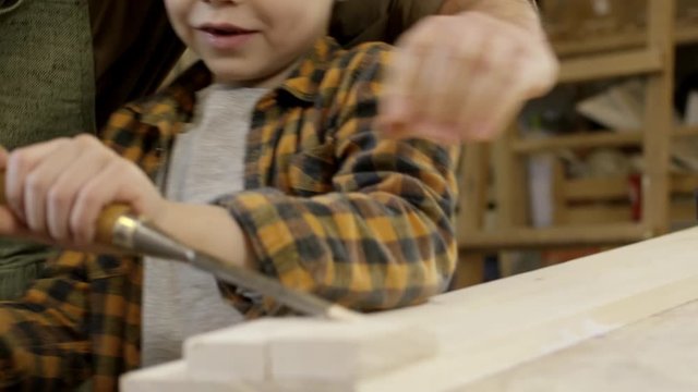 Tilt down of cheerful father teaching little son to carve wood with chisel and mallet while spending time together in carpentry workshop