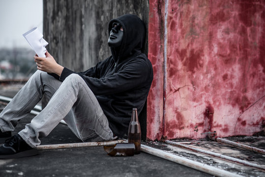 Mystery man wearing black mask and hoody jacket holding white mask sitting on rooftop of abandoned building, depression self destruction suicidal addiction massive depressive disorder concept
