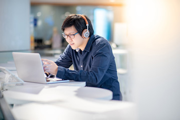 Young Asian business man listening to music by headphones and smartphone while working with laptop computer in co working space. freelance or digital nomad lifestyle concepts