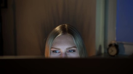 Woman peeking out computer monitor at night. Crop view of beautiful female sitting at night at computer and looking at camera over top of monitor with dark window on blurred background