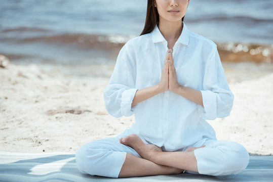 Cropped image of woman meditating in anjali mudra (salutation seal) pose on yoga mat by sea