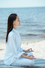 side view of young asian woman in ardha padmasana (half lotus pose) on yoga mat by sea © LIGHTFIELD STUDIOS