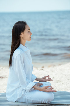 side view of young asian woman in ardha padmasana (half lotus pose) on yoga mat by sea
