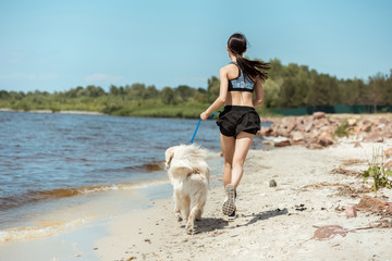 rear view of female jogger running with dog on beach near sea