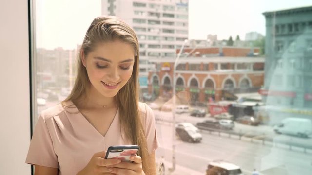 Young beautiful girl is sitting on window, chating on smartphone in daytime, communication concept, urban concept
