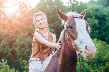 People and animals friendship, hippotherapy concept. Portrait of happy pensive woman cowgirl, caress a brown horse while riding. Clothed white jeans shorts, brown leather vest. Has slim sport body.