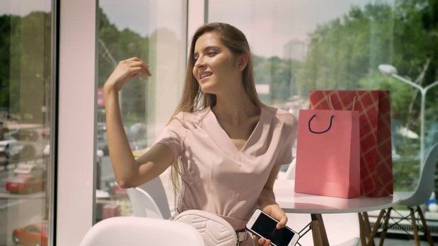 Young beautiful girl is sitting in cafe with big windows in daytime, touching hair, holding smartphone, bags on table