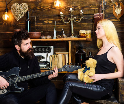 Romantic date concept. Couple in love on relaxed faces enjoy romantic atmosphere. Man play guitar while lady holds teddy bear in hands. Couple spend romantic evening, wooden interior background
