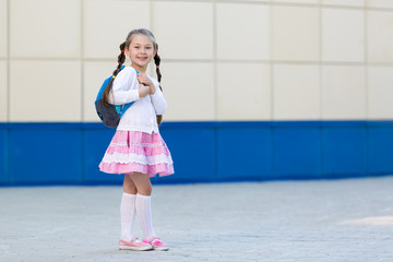 The girl is a young fashionista posing. A little beautiful girl in a summer dress with a backpack is standing by the wall in the city.