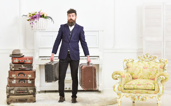 Macho attractive, elegant on surprised face carries vintage suitcases. Man with beard and mustache wearing classic suit delivers luggage, luxury white interior background. Butler and service concept.