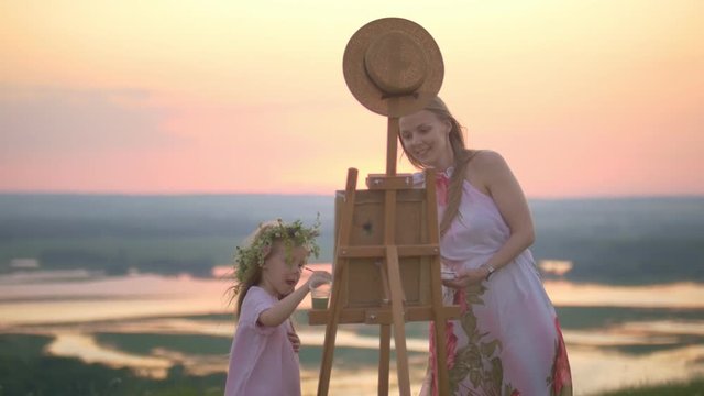 Young caucasian woman with daughter painting nature on the summer hill at sunset