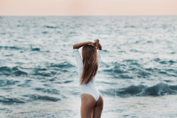 Beautiful young woman with dark hair in white swimsuit is posing on the beach on the background of azure sea waves. Summer day near ocean. Copy space