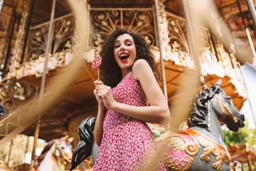 Young joyful lady with dark curly hair in dress standing with lolly pop candy in hands and happily looking in camera with beautiful carousel on background