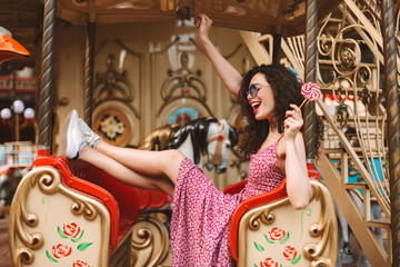 Cheerful lady with dark curly hair in sunglasses and dress holding lolly pop candy in hand and...
