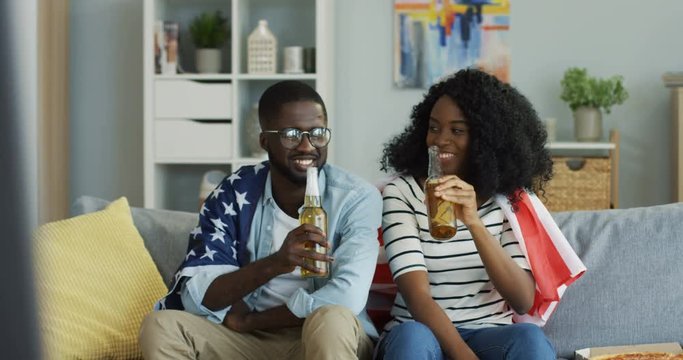 African American young happy couple doing cheers gesture and drinking beer while sitting on the sofa under the American flag in the living room. Indoors.