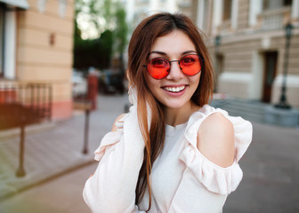 Portrait Happy young adult woman smiling with teeth smile and long brunette hair outdoors ,walking on city street at sunset time wearing trendy sunglasses,beige shirt and stylish bag.Toned,copy space.