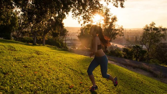 The sun rises behind a fit woman working out by running up a grass covered hill.