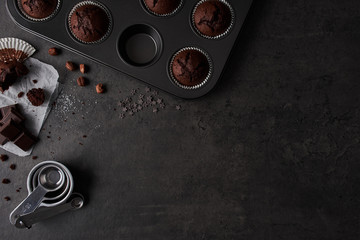 Baking background with chocolate muffins in bakeware, measuring cups, chocolate, sugar and nuts on dark concrete background. Flat lay with copy space.
