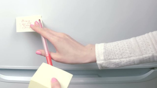 Closeup woman's hands stick yellow sticker paper sheets with words buy: milk, bread, chicken. Family note.