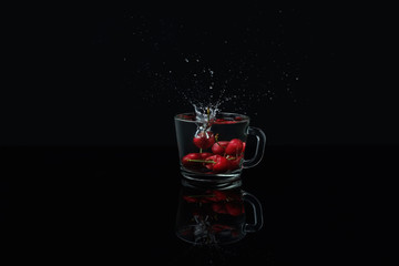 Macro photo of a clear glass mug with cherries in front of a black background on a black table made of glass. Reflection of a glass mug with cherries. Falling berries create splashes of water.