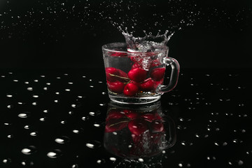 Clear glass mug with cherries in front of a black background on a black table made of glass covered with water drops. Reflection of a glass mug with cherries. Falling berries create splashes of water.