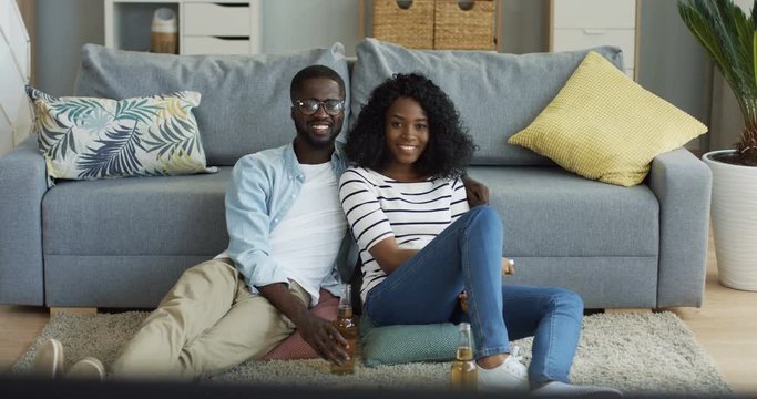 Young happy African American couple hugging while sitting on the floor in the living room and watching TV, man drinking beer. Inside.