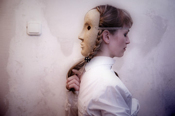 Blonde girl in a gray theatrical mask stands against the wall