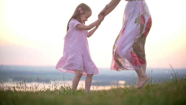 Cute little girl holding hands with mom stomps on the grass hill at sunset