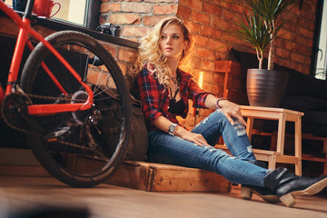 Sensual blonde hipster girl with long curly hair dressed in a fleece shirt and jeans sitting on a wooden box, looking away, at a studio with loft interior.