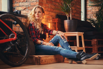 Fototapeta na wymiar Sensual blonde hipster girl with long curly hair dressed in a fleece shirt and jeans sitting on a wooden box, looking away, at a studio with loft interior.