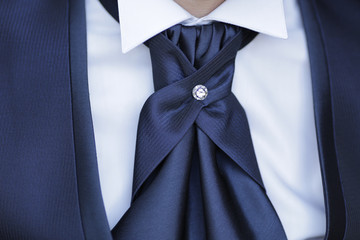Dettaglio abito da sposo blu con camicia bianca, giacca blu, cravatta nera con gioiello al centro