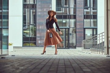 Heureuse femme élégante de mode portant une veste noire, un chapeau marron et une jupe avec une pochette de sac à main marchant sur le centre-ville européen.