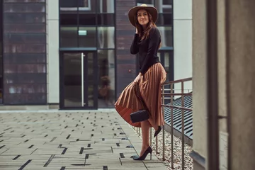 Gordijnen Glimlachende mode elegante vrouw draagt een zwarte jas, bruine hoed en rok met een handtas clutch poseren terwijl leunend op een stalen reling op het Europese stadscentrum. © Fxquadro