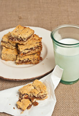 Top view, medium distance of a fig almond bar on a paper napkin, a glass of milk, five bars on a round, white plate with brown, rim on a burlap place mat