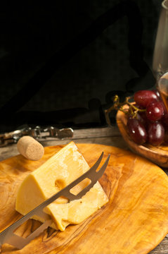 Top view, medium distance of a wedge of cheese with a cheese knife cutting on a rare, wood plate with a round, wood bowl of freshly picked red grapes  and a corkscrew and cork, wine tasting event