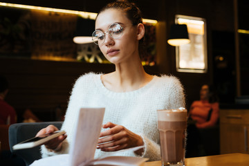 beautiful young girl student in glasses and a white sweater sits in a cafe after studying at university