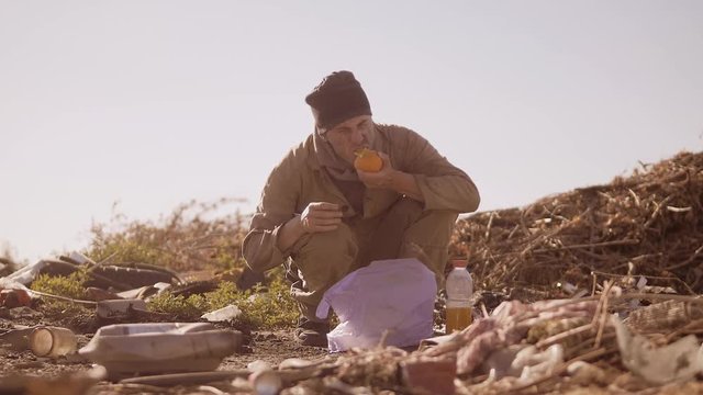 portrait of a dirty homeless hungry man in a dump eating orange for food in the package with walking goes looking for food slow motion video. homeless dirty man roofless person looking for food in a