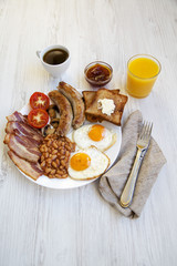 Full English breakfast with fried eggs, bacon, sausages, beans and toasts. White wooden surface.