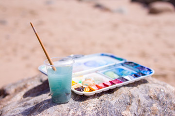 Artistic equipment: paint brushes, tubes of paint, palette and paintings on rock in nature sea shore at sunny summer day - creation, drawing and freedom concept