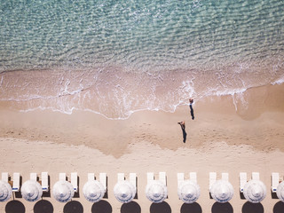 Aerial view of amazing turquoise sea with white umbrellas and sun loungers. Two people are walking on the shore. Beautiful sunny summer day in Sardinia, Mediterranean sea, Italy..