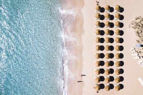 Aerial View Of Amazing Turquoise Sea With Brown Straw Umbrellas And Sun Loungers. Two People Are Walking On The Shore. Beautiful Sunny Summer Day In Sardinia, Mediterranean Sea, Italy..